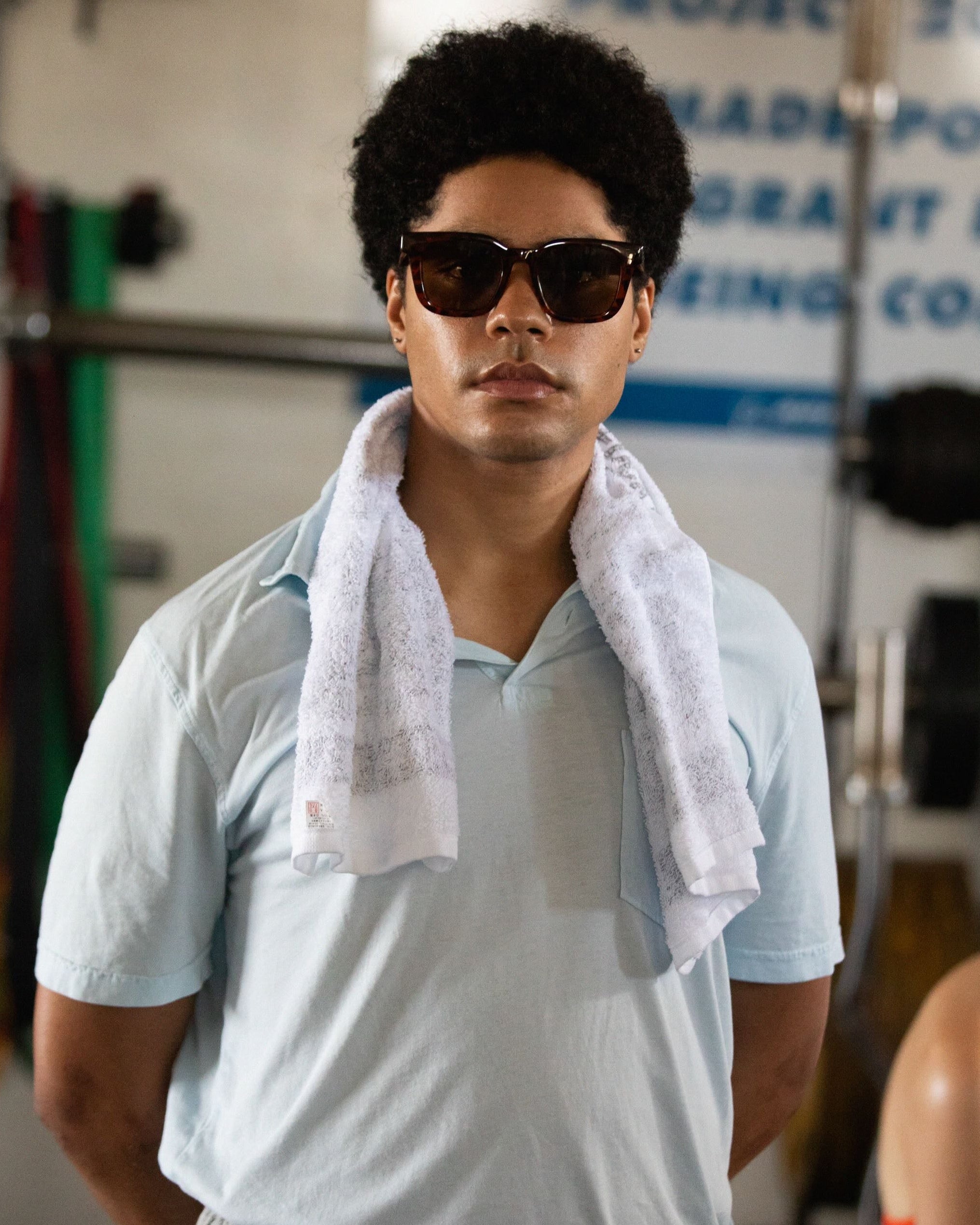 Person wearing sunglasses and a light blue shirt with a towel draped over their shoulder in a gym setting.